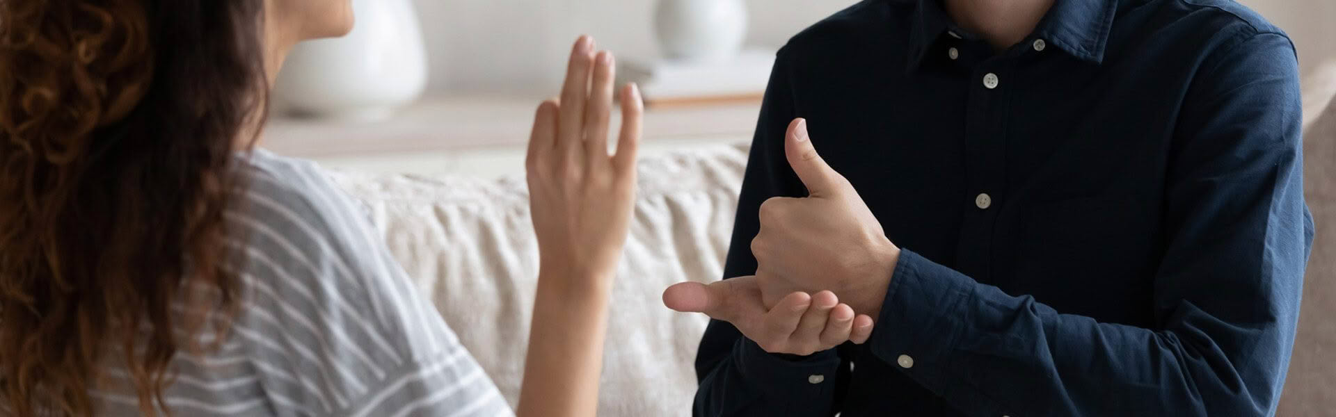 A woman in a grey t shirt is sitting in front of a man wearing a blue shirt, she is signing OK in NZSL, the man is signing KIA ORA in NZSL to her