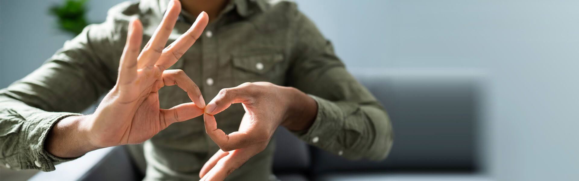 A man wearing a light green shirt is sitting on a couch, he is signing INTERPRETER in NZSL close to the camera