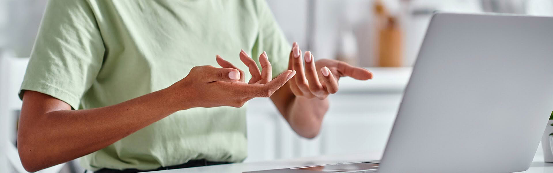A woman wearing a light green t shirt is sitting in front of a laptop computer, she is signing DISCUSSION in NZSL to someone on the computer screen